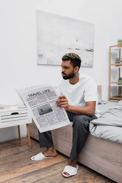 Serious african american man with dyed hair sitting on bed and reading travel life newspaper — Stock Photo