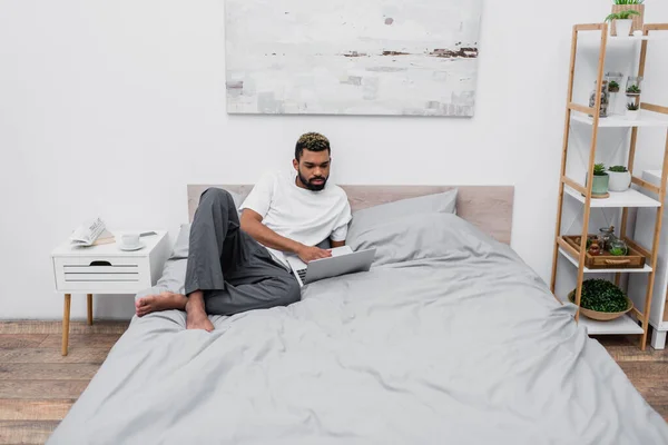 Hombre afroamericano con el pelo teñido usando el ordenador portátil y trabajando desde casa en la cama - foto de stock