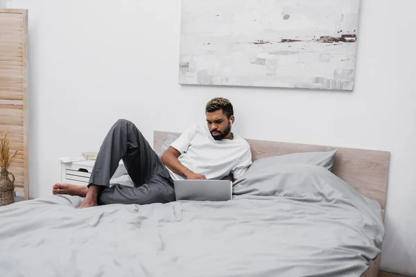 Афроамериканський чоловік в бездротових навушниках використовує ноутбук і працює з дому в ліжку — стокове фото