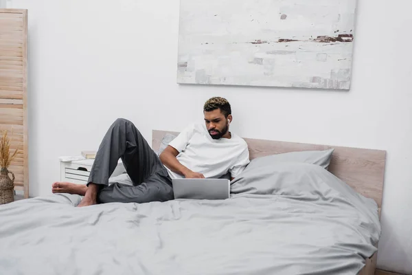 Hombre afroamericano en auriculares inalámbricos utilizando el ordenador portátil y trabajando desde casa en la cama - foto de stock