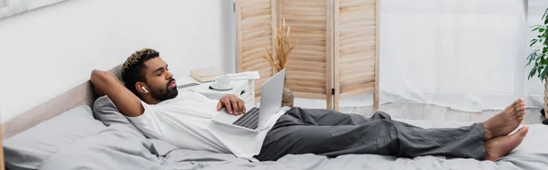 Афроамериканський чоловік у бездротових навушниках дивиться фільм, лежачи на ліжку з ноутбуком, банер — стокове фото
