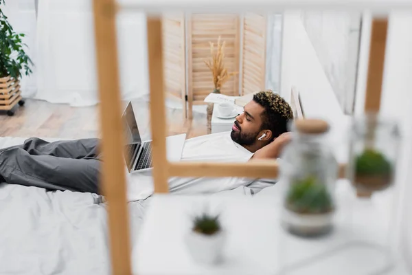 Африканский американец в беспроводных наушниках смотрит кино, лежа на кровати с ноутбуком рядом с размытой стойкой — стоковое фото