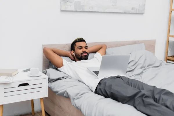Hombre afroamericano feliz en auriculares inalámbricos viendo la película mientras está acostado en la cama con el ordenador portátil - foto de stock