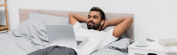 Hombre afroamericano feliz en auriculares inalámbricos viendo la película mientras está acostado en la cama con el ordenador portátil, pancarta - foto de stock