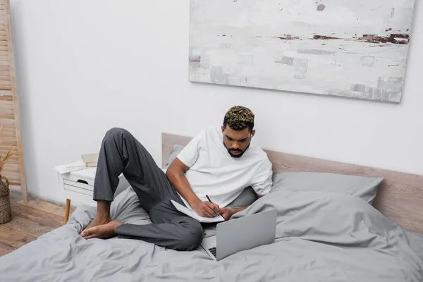 African american man in wireless earphones using laptop and writing in notebook in bedroom — Stock Photo