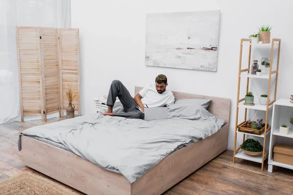 Uomo afroamericano barbuto in auricolare senza fili utilizzando il computer portatile e lavorando da casa a letto — Foto stock