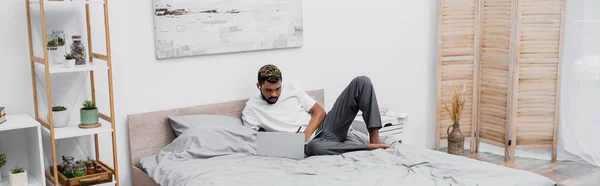 Homem americano africano barbudo em fone de ouvido sem fio usando laptop e trabalhando de casa na cama, banner — Fotografia de Stock