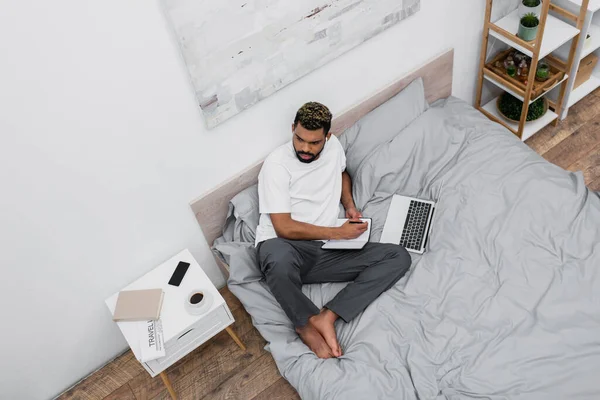 Вид зверху на афроамериканця, який використовує ноутбук і працює з дому в ліжку — стокове фото