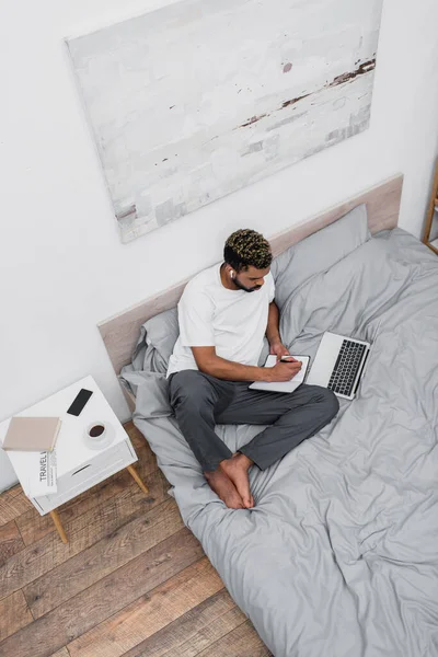 Vista superior del hombre afroamericano en auriculares inalámbricos utilizando el ordenador portátil y trabajando desde casa en la cama - foto de stock