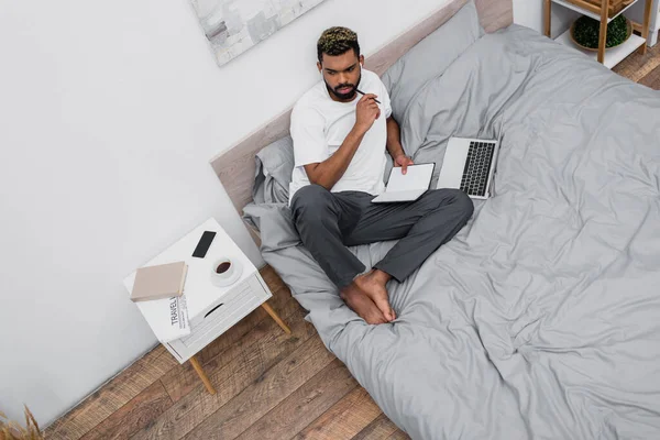 Vista superior do homem americano africano pensativo em fones de ouvido sem fio usando laptop e trabalhando de casa na cama — Fotografia de Stock