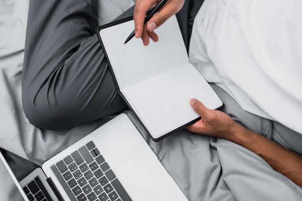 Vista ritagliata dell'uomo afro-americano con notebook e penna vicino al computer portatile moderno — Foto stock