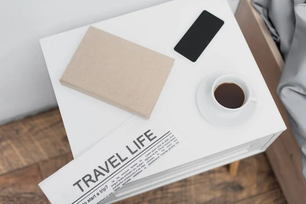 Top view of cup with coffee and smartphone near newspaper with travel life lettering and book on white bedside table — Stock Photo
