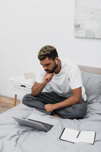 Thoughtful african american man in wireless earphone using laptop and working from home in bed — Stock Photo