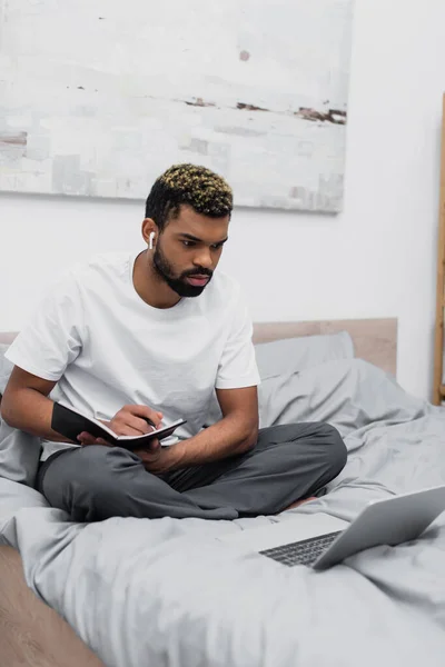 Focused african american man in wireless earphone using laptop and holding notebook in bed — Stock Photo