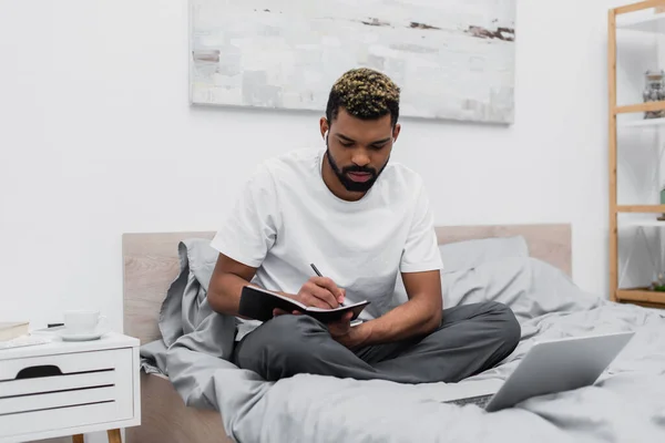Bearded african american man in wireless earphones using laptop and writing in notebook — Stock Photo