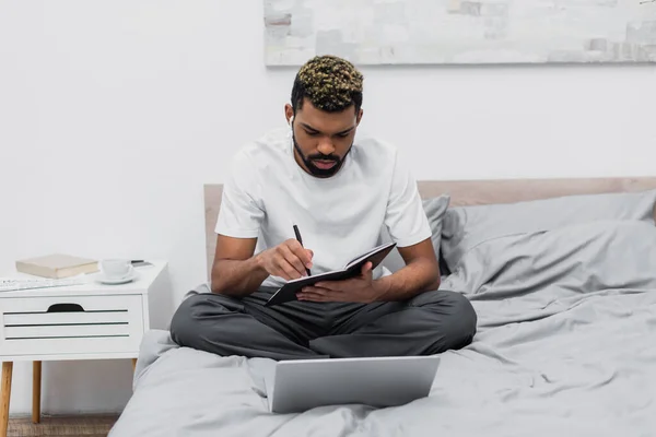 Бородатий афроамериканський чоловік в бездротових навушниках дивиться на ноутбук біля ноутбука на ліжку — стокове фото