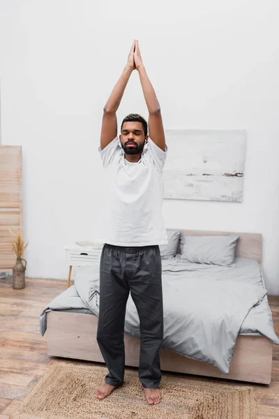 Barbudo africano americano hombre con los ojos cerrados de pie en árbol yoga pose en dormitorio - foto de stock