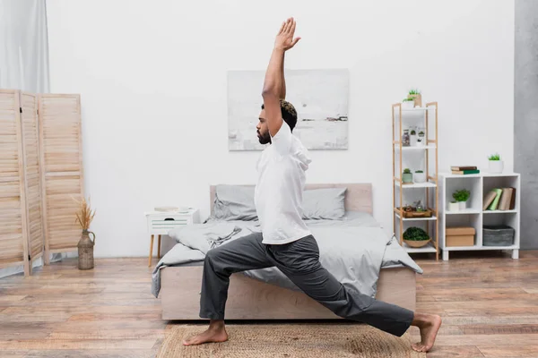 Vista lateral del hombre afroamericano barbudo practicando yoga en pose guerrera y meditando en el dormitorio - foto de stock