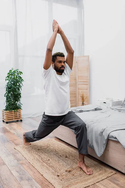 Uomo afroamericano barbuto praticare yoga in posa guerriero e meditare in camera da letto moderna — Foto stock