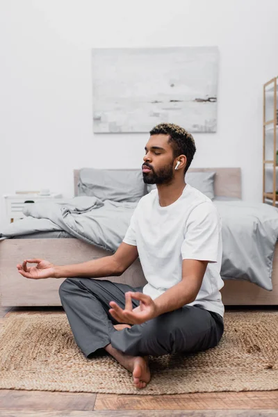 Bearded african american man in wireless earphone meditating with closed eyes while sitting near bed at home — Stock Photo