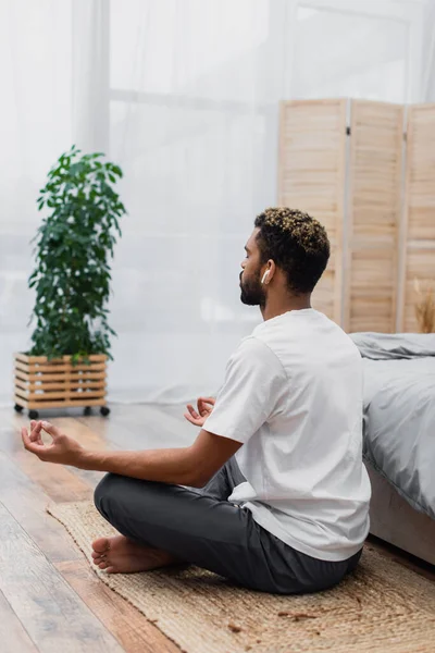 Homem americano africano barbudo em fone de ouvido sem fio meditando enquanto sentado perto da cama em casa — Fotografia de Stock