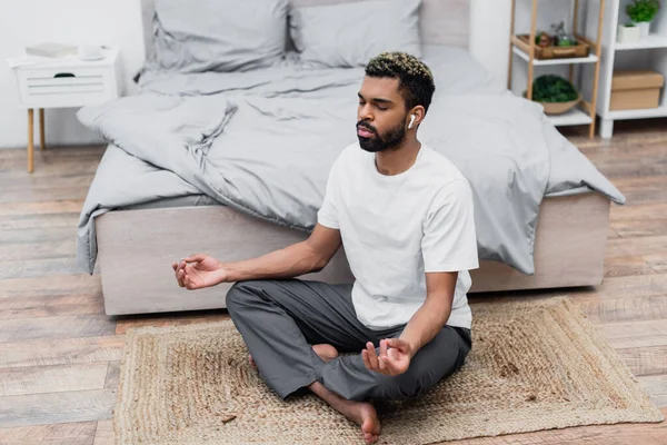 Barbudo afroamericano hombre meditando en la alfombra mientras está sentado cerca de la cama en casa - foto de stock