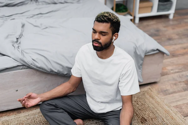 Vue grand angle de barbu homme afro-américain méditant avec les yeux fermés tout en étant assis près du lit à la maison — Photo de stock