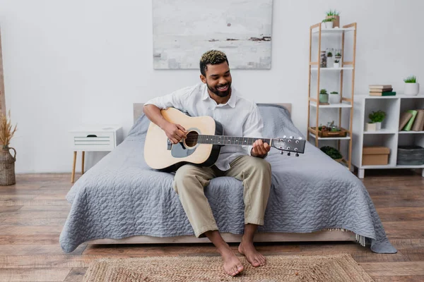 Descalzo y feliz afroamericano hombre con el pelo teñido y la barba tocando la guitarra acústica en el dormitorio - foto de stock