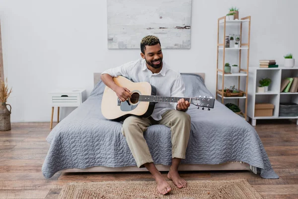 Barefoot and happy african american man with dyed hair and beard playing acoustic guitar in bedroom — Stock Photo