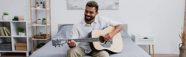 Pleased african american man with dyed hair and beard playing acoustic guitar in bedroom, banner — Stock Photo