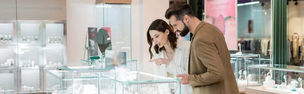 Mujer morena señalando joyas en escaparate cerca de novio en la tienda, pancarta - foto de stock