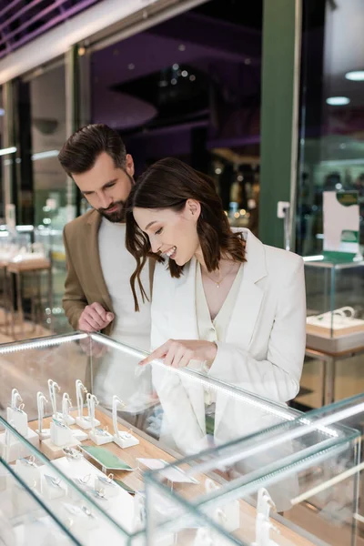 Mujer joven apuntando a la joyería en escaparate cerca de novio en la tienda - foto de stock