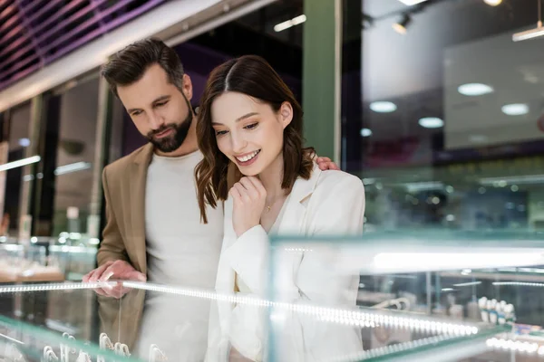 Man hugging smiling girlfriend near blurred showcase in jewelry shop - foto de stock