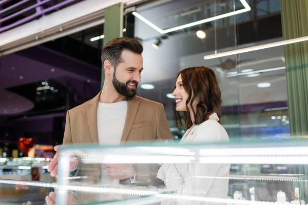 Smiling man pointing at showcase and looking at girlfriend in jewelry shop — Stock Photo