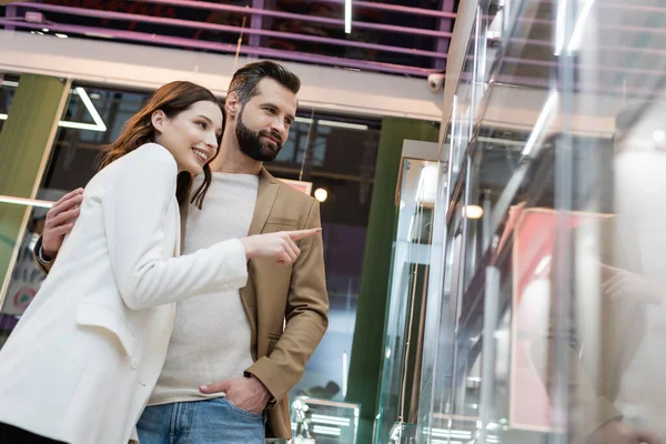 Низький кут зору чоловіка обіймає усміхнену дівчину біля вітрини ювелірного магазину — стокове фото
