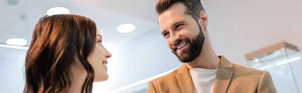 Бородатый мужчина смотрит на девушку брюнетку в размытом ювелирном магазине, баннер — стоковое фото