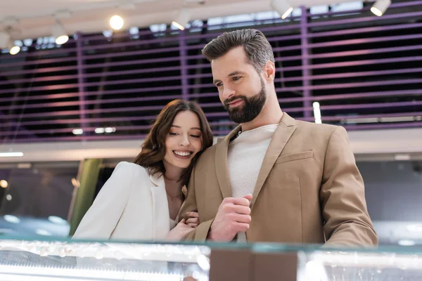 Mujer sonriente abrazando novio cerca de escaparate borroso en joyería — Stock Photo