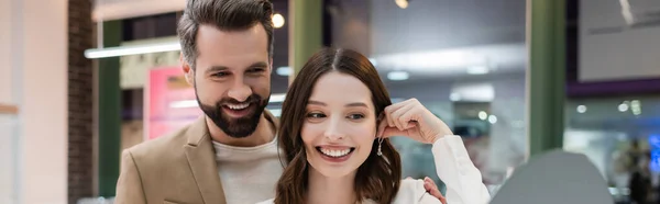 Donna sorridente che tiene l'orecchino vicino allo specchio e al fidanzato in gioielleria, banner — Foto stock