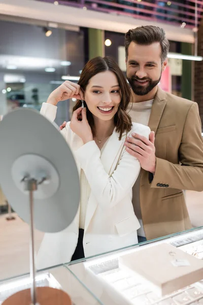 Cheerful man hugging girlfriend wearing earring near mirror in jewelry shop — Stock Photo