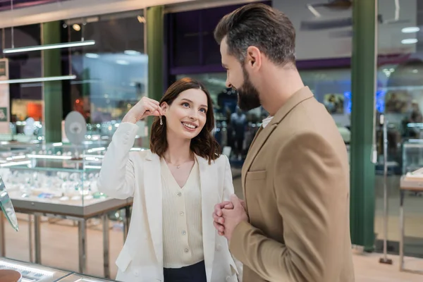 Femme positive tenant boucle d'oreille près du petit ami et miroir dans la bijouterie — Photo de stock