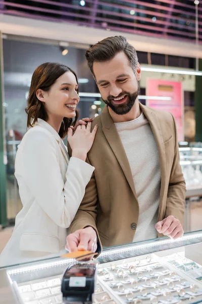 Sorrindo mulher abraçando namorado pagando com cartão de crédito na joalharia — Fotografia de Stock