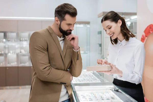 Vendedor sonriente apuntando a la joyería cerca de cliente enfocado en tienda - foto de stock