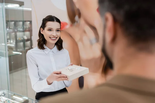 Venditore sorridente che tiene gioielli sulla scatola vicino al cliente offuscato in negozio — Foto stock