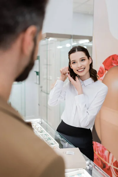 Smiling seller holding jewelry near blurred man in store — Stockfoto