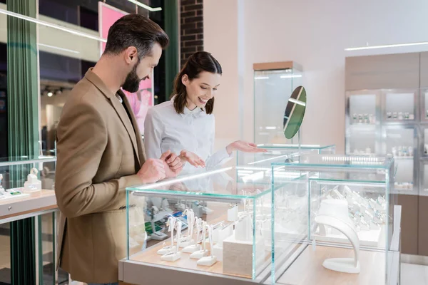 Vendedor sonriente señalando escaparate cerca del espejo y el cliente en la joyería - foto de stock