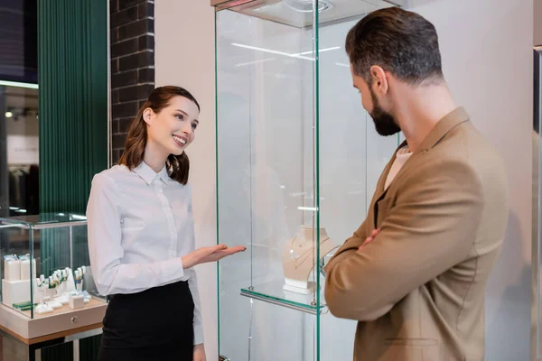 Venditore sorridente che indica collane in vetrina vicino al cliente in gioielleria — Foto stock