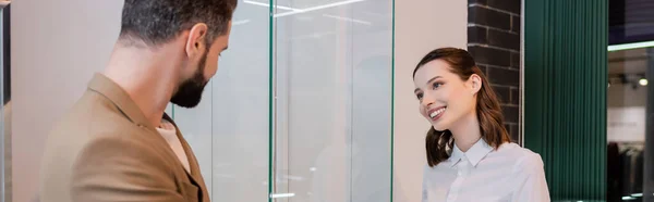 Vendedor positivo mirando al cliente borroso cerca de escaparate de cristal en la joyería, pancarta - foto de stock