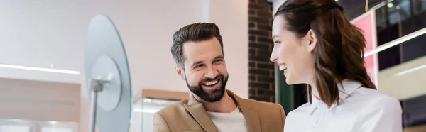 Vendedor sonriente y cliente mirándose el uno al otro cerca del espejo borroso en la joyería, pancarta - foto de stock