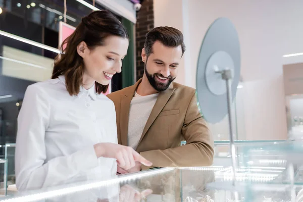 Smiling client looking at showcase near seller and blurred mirror in jewelry shop — Photo de stock