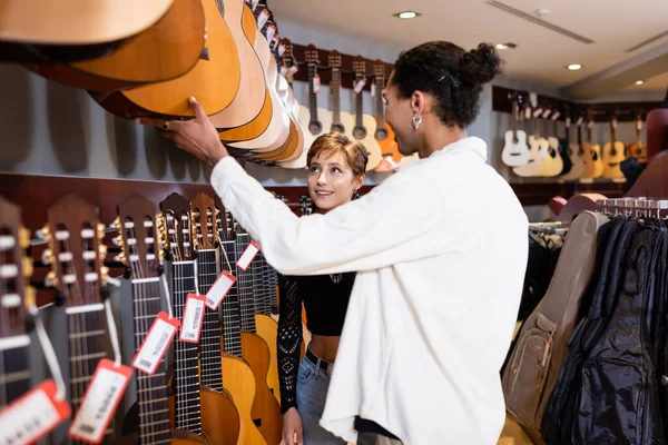 Улыбающийся покупатель смотрит на акустическую гитару рядом с африканским продавцом в музыкальном магазине — стоковое фото